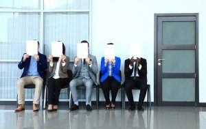 Five people are sat in a row, on chairs, in a corridor. Each of them is wearing a suit and their faces are covered by a piece of white card theat they each hold over their own faces.