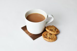 A white cup on a wooden coaster contains tea with milk. To the side of the cup lie two chocolate chip cookies.